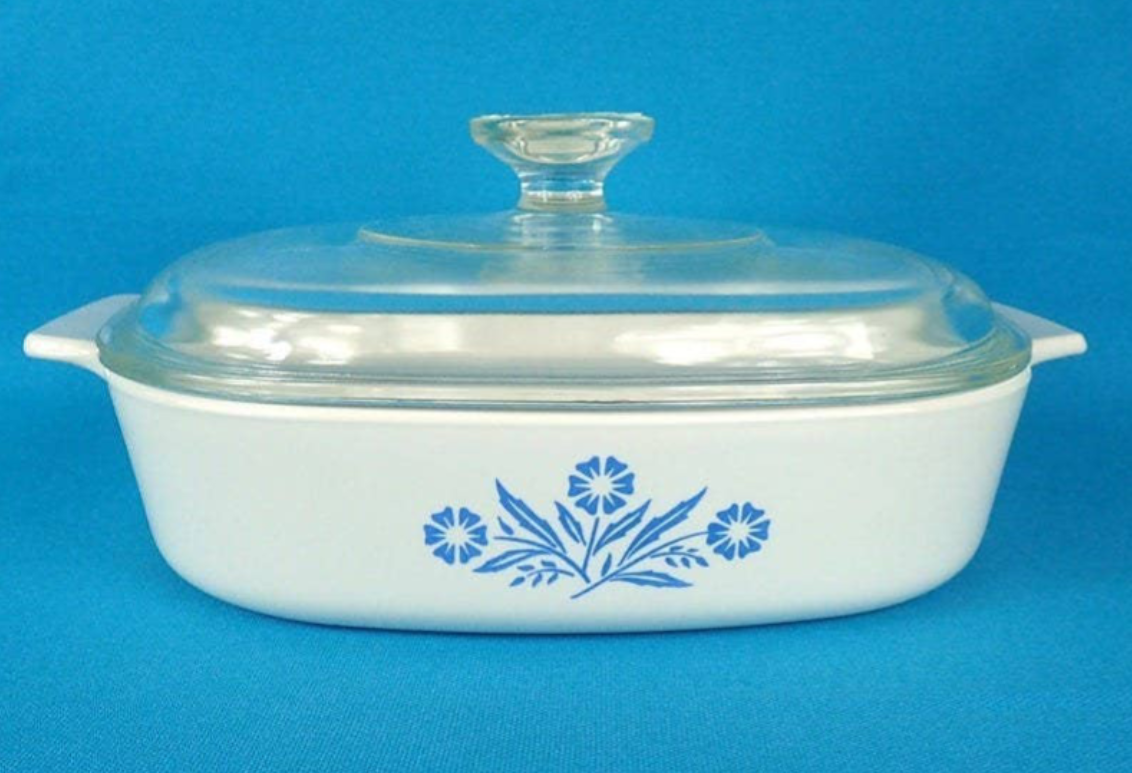 A white casserole dish with handles and a clear glass lid, featuring a blue floral design on the front, set against a solid blue background.