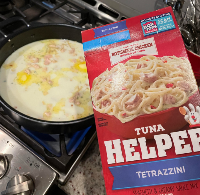 A hand holds a Tuna Helper Tetrazzini box near a stove with a skillet containing a creamy mixture. The box mentions using rotisserie chicken instead of tuna.