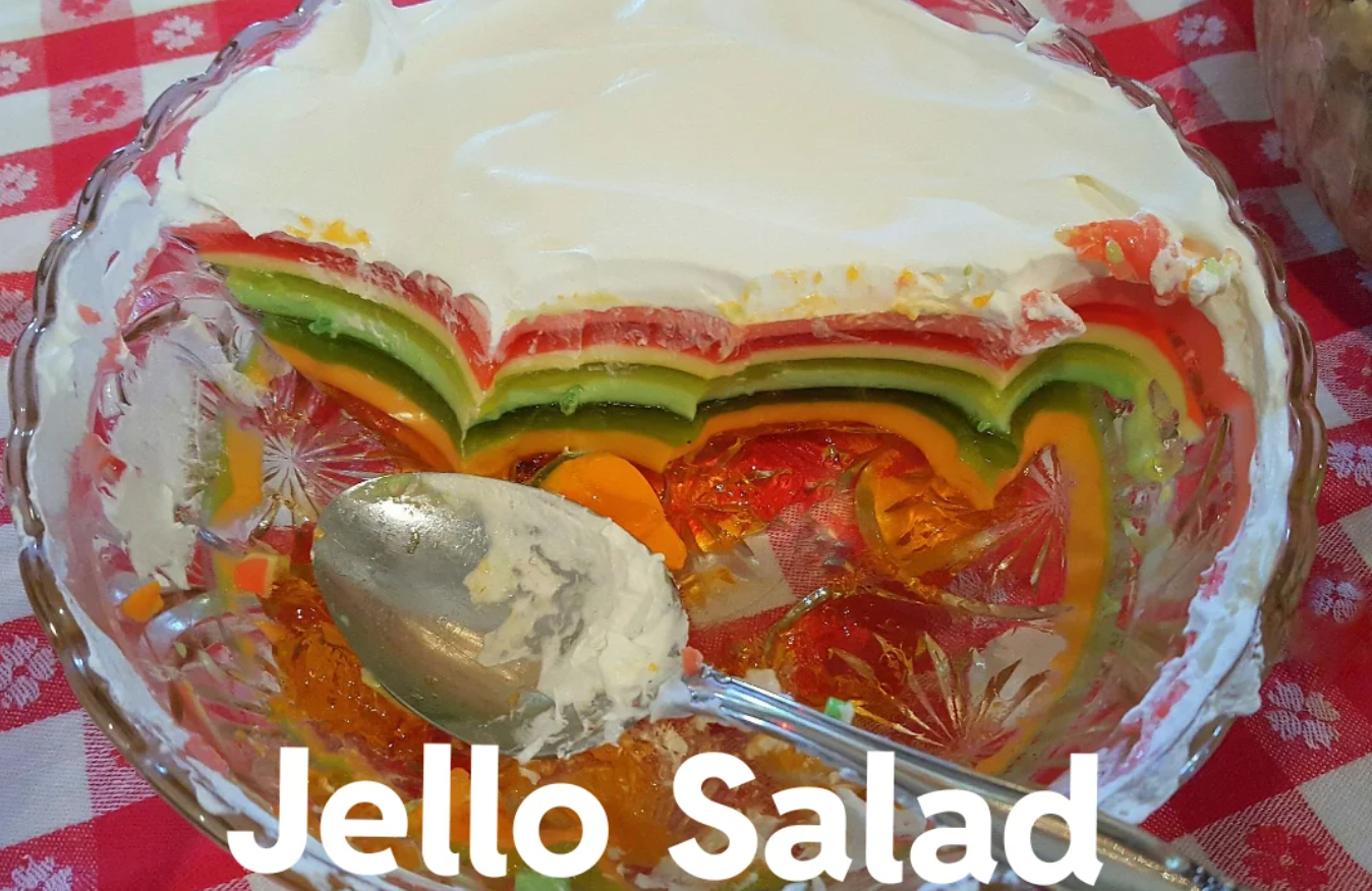 A glass bowl of colorful layered Jello salad with whipped topping, partially eaten, sits on a red checkered tablecloth. A spoon with whipped topping rests in the bowl. The text "Jello Salad" appears at the bottom.
