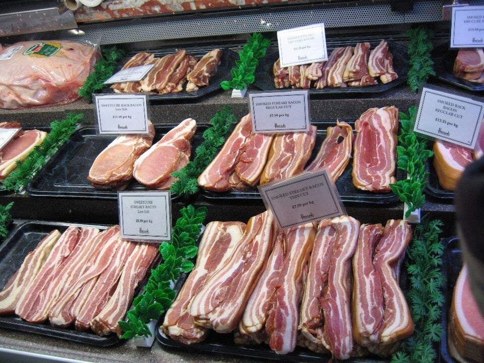 Trays of sliced raw bacon are displayed at a butcher’s shop counter, separated by price labels and garnished with green decorative parsley.