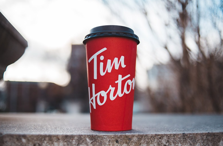 A red Tim Hortons coffee cup with a black lid sits on a concrete surface outdoors, representing one of the most iconic coffee houses. The cup features the brand's white cursive logo, with blurred trees and buildings in the background.