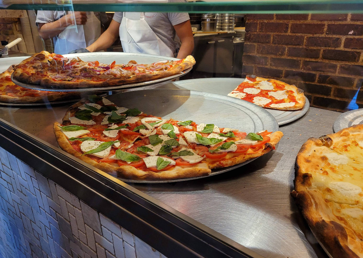Several large pizzas with various toppings, including fresh basil, cheese, and tomato, are displayed on metal trays behind a glass counter—perfect if you’re after a quality slice of pizza for cheap, while chefs in aprons prepare food in the background.