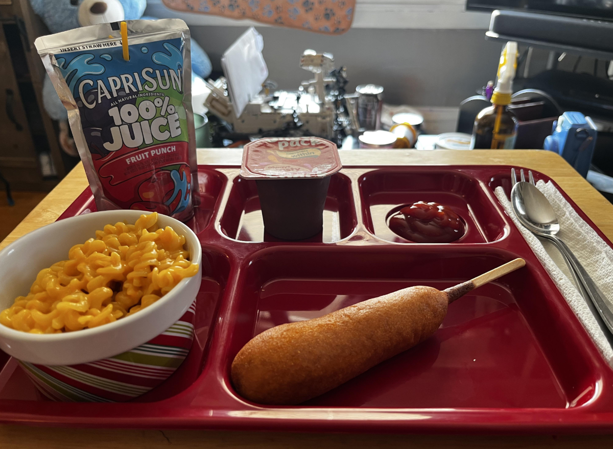 A red tray holds macaroni and cheese, a corn dog, ketchup, chocolate pudding, and a Capri Sun juice pouch on a wooden table in a room with various items in the background.