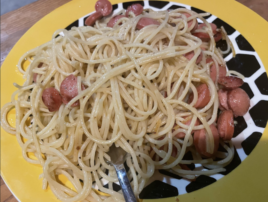 A plate of spaghetti mixed with sliced sausages, served on a yellow and black patterned plate with a fork resting on the side.