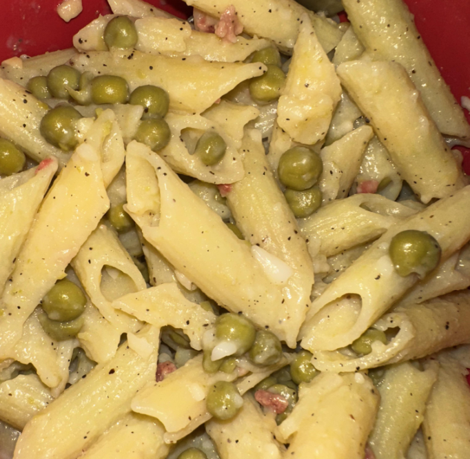 Close-up of cooked penne pasta mixed with green peas and small bits of bacon in a creamy sauce, seasoned with black pepper.