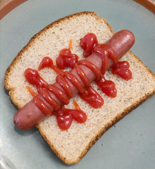 A hot dog on a slice of bread with ketchup squirted in a zigzag and heart shapes on top.