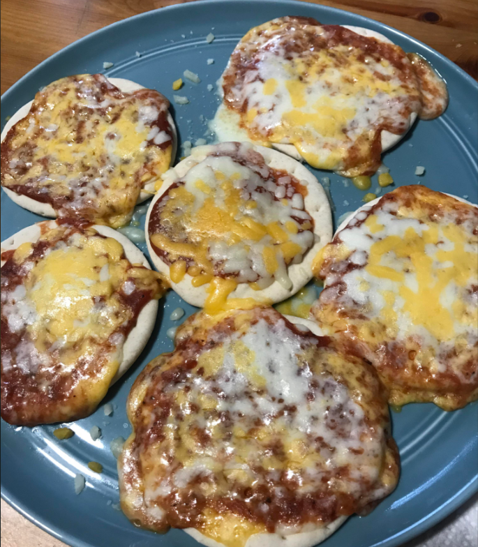 Six small homemade pizzas covered in melted cheese and tomato sauce are arranged on a blue plate, with some cheese spilling over the edges of the crusts.