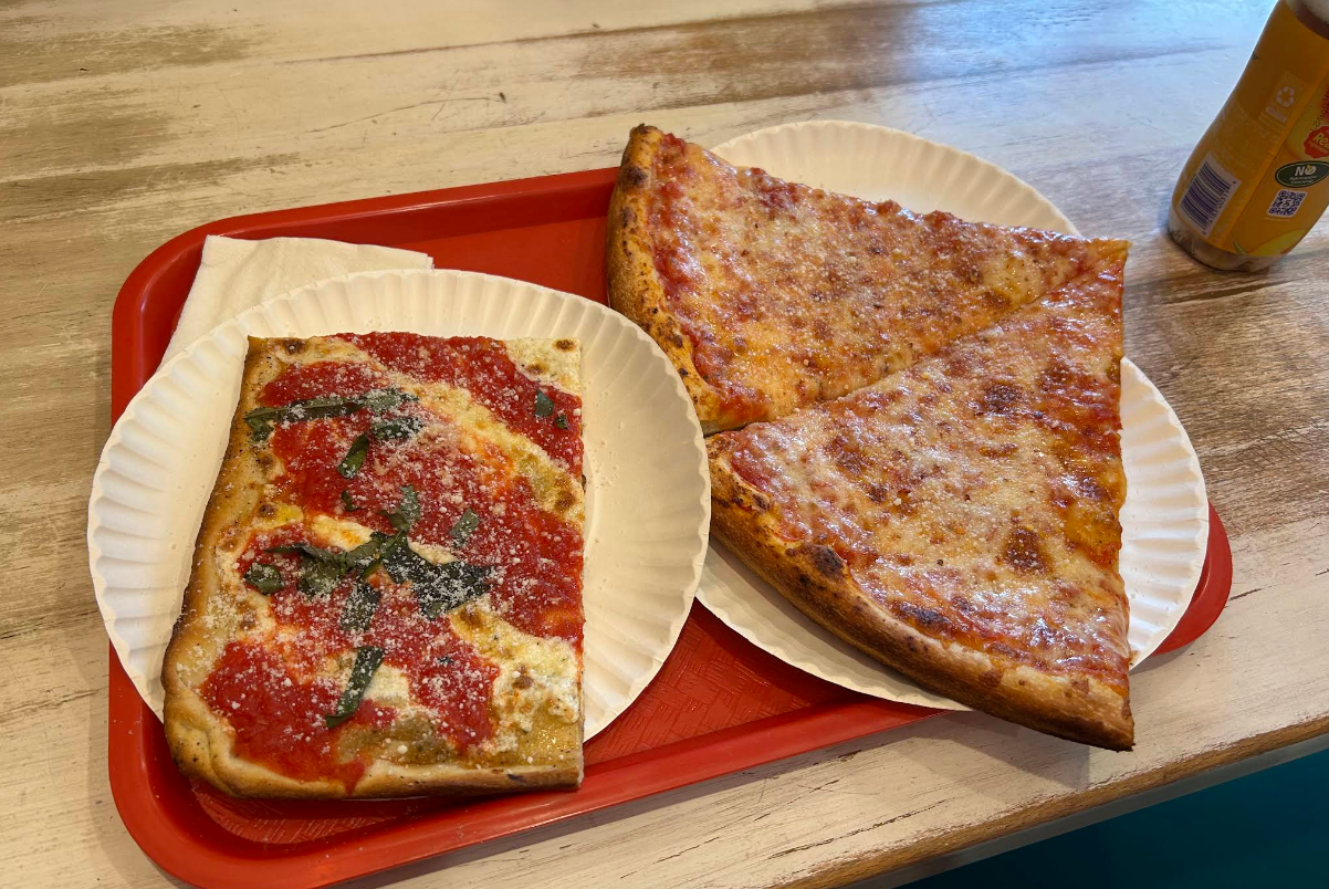 A red tray holds two paper plates: one with a square slice of pizza topped with tomato sauce, cheese, and basil, the other with two large triangular slices of cheesy pizza—a quality slice of pizza for cheap. A canned drink sits in the corner.
