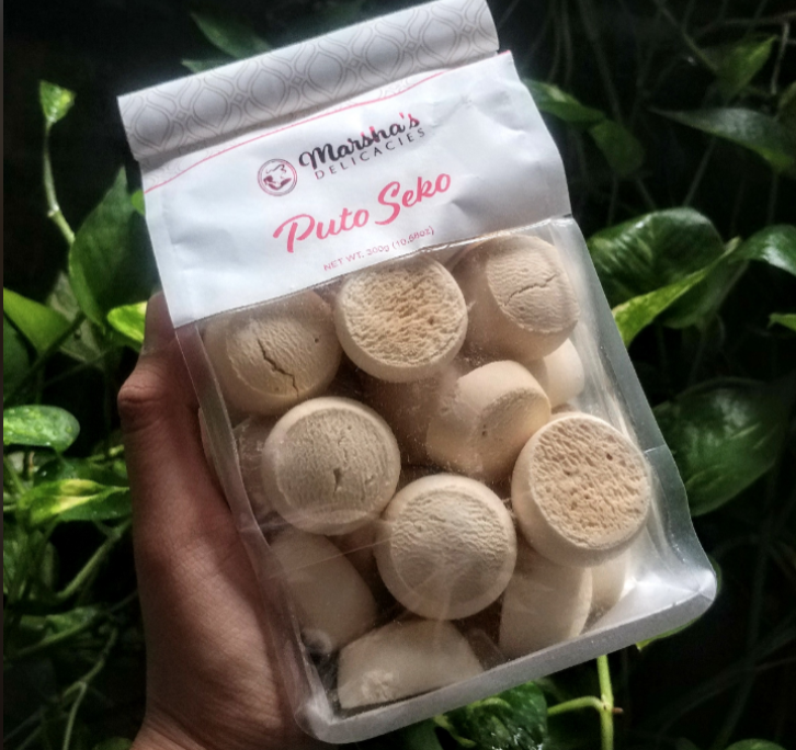 A hand holds a clear package labeled "Marsha's Delicacies Puto Seko," filled with round, beige Filipino rice cookies, against a leafy green background.