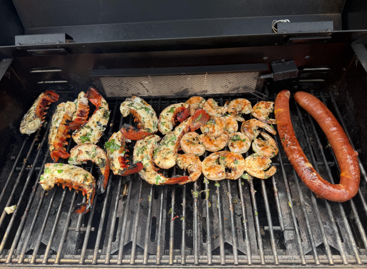 Lobster tails, shrimp, and a sausage cooking on a barbecue grill. The seafood is seasoned and arranged in the center, with the sausage on the right side of the grill.