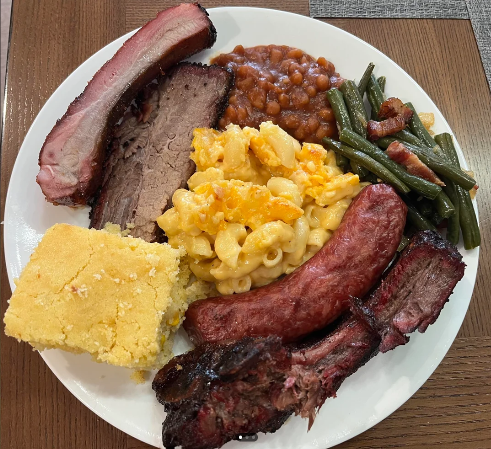 A plate with barbecue ribs, sausage, brisket, macaroni and cheese, baked beans, green beans with bacon, and a slice of cornbread on a wooden table.