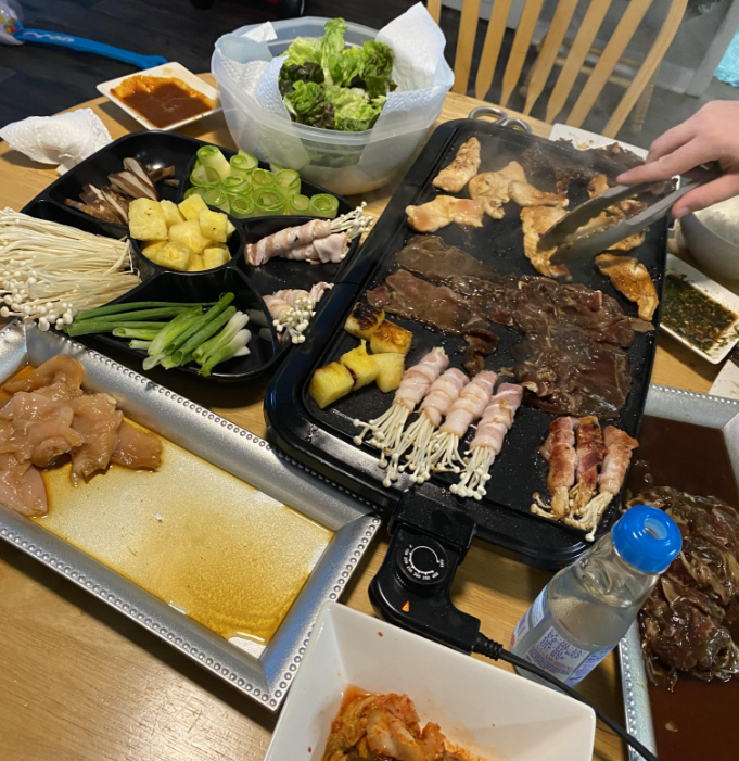 A tabletop grill with assorted meats, mushrooms, and vegetables cooking, surrounded by raw ingredients like chicken, greens, and sauces. A hand holds tongs over the grill.