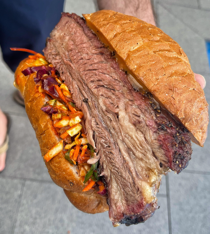A hand holds a sandwich with thick, juicy beef brisket and colorful slaw in a golden bun, with gray pavement in the background.