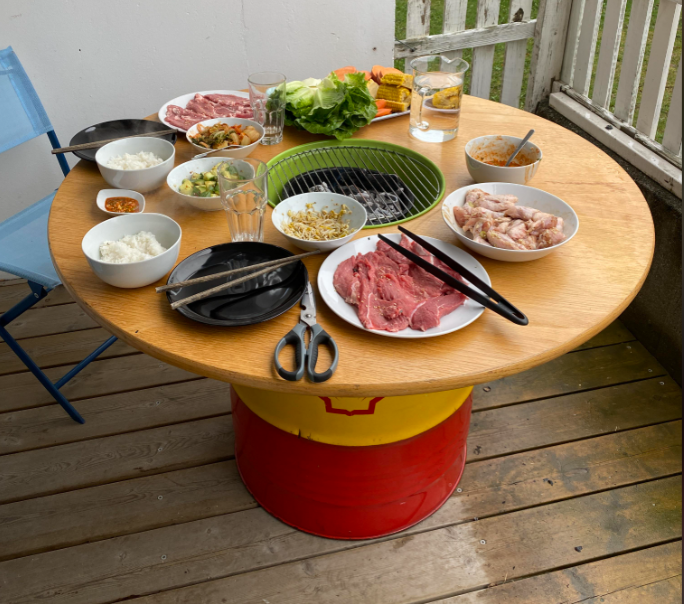 Round table with a grill in the center, surrounded by bowls of meat, rice, vegetables, tongs, plates, scissors, and drinks. The table stands on a red and yellow drum on a wooden deck. Blue chair visible on the left.