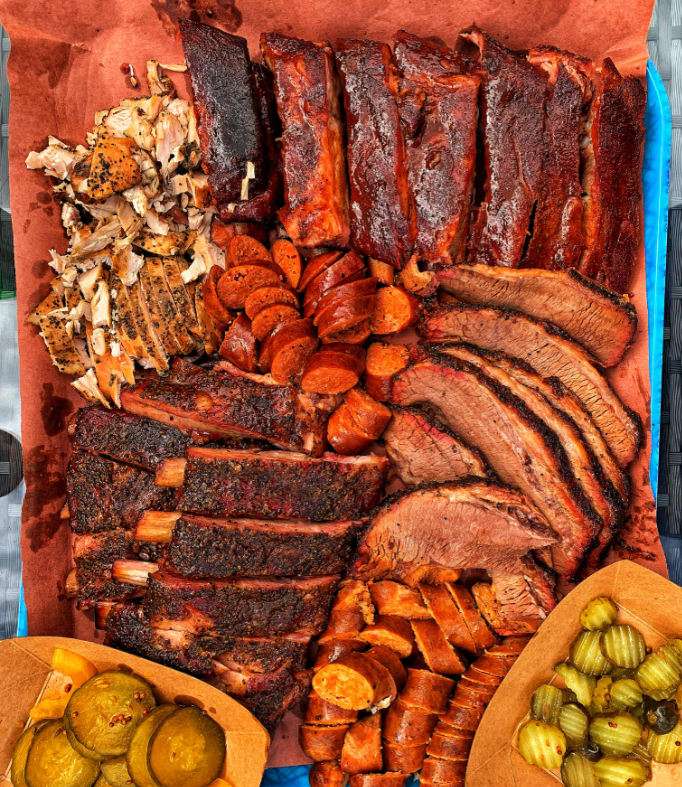 A tray of assorted smoked barbecue meats, including ribs, brisket slices, sausage, and pulled chicken, with sides of sliced pickles and jalapeños in separate brown paper containers.