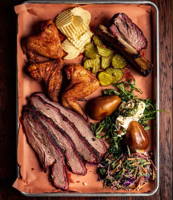 A tray of barbecue foods: sliced brisket, smoked chicken wings, ribs, potato chips, pickles, two whole pears, salad with arugula and slaw, served on brown paper over a metal tray.