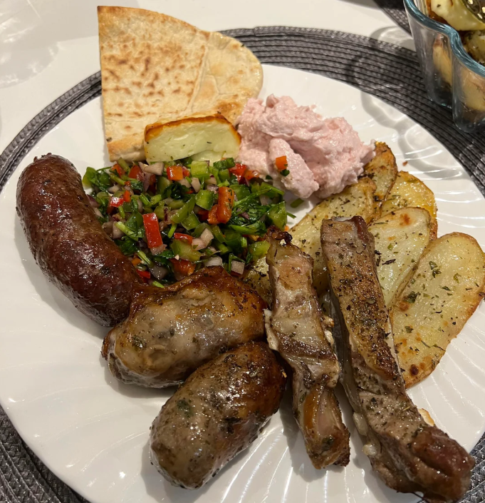 A white plate with grilled sausages, lamb chops, potato wedges, a green salad with tomatoes, pita bread, a slice of halloumi cheese, and a pink spread, all on a woven placemat.