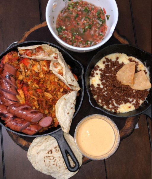 A wooden tray holds tortillas, grilled sausage, chicken fajitas, salsa, melted cheese with chorizo, and a small bowl of queso dip, all in cast iron skillets and bowls.
