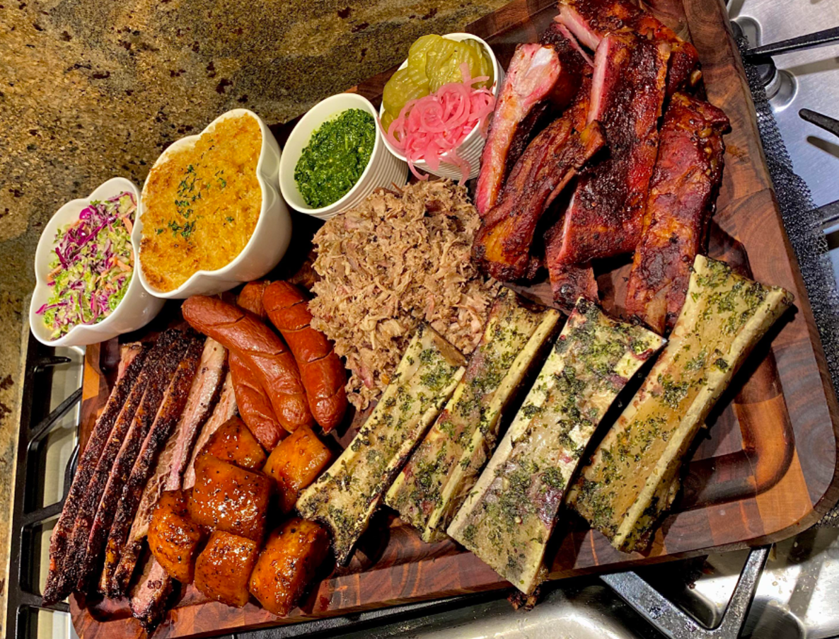 A wooden board filled with barbecue ribs, sausages, brisket, herbed bone marrow, rice, pickles, coleslaw, sweet potatoes, pickled onions, sauce, and cornbread. The platter is set on a kitchen counter.