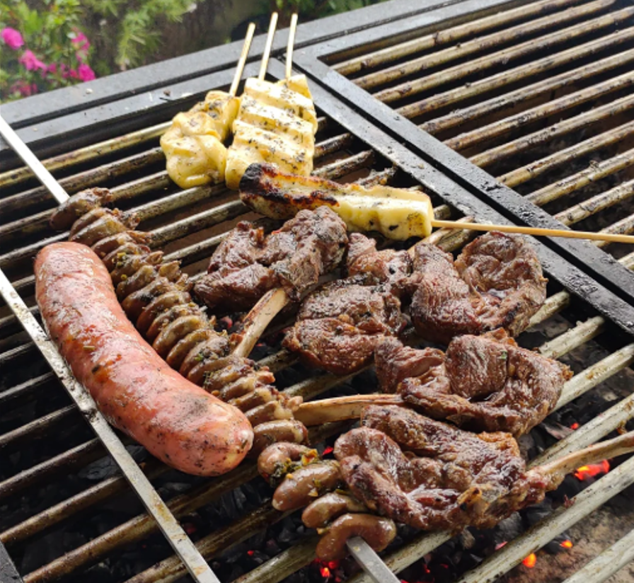 Various meats, including beef, sausage, and chicken skewers, are being grilled over an open barbecue. The grill has visible glowing coals and a few pink flowers are seen in the background.