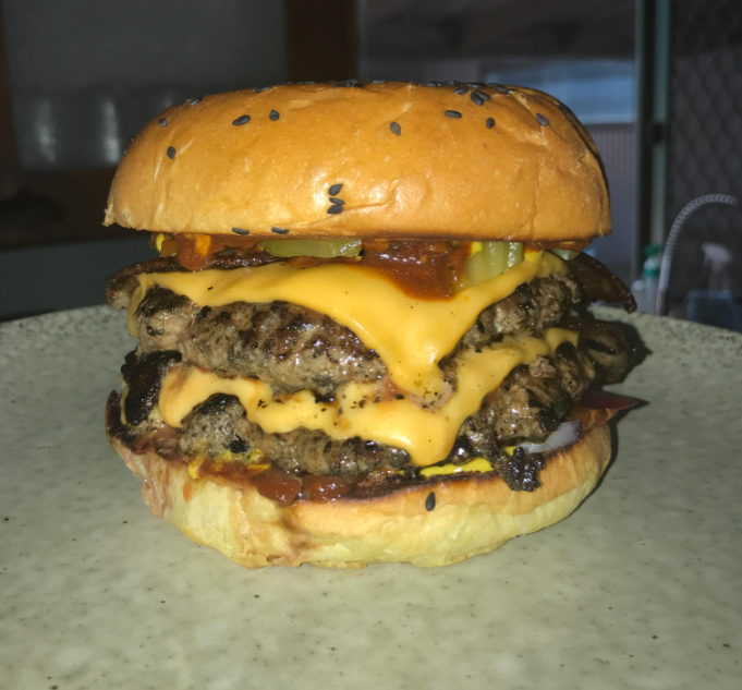 A cheeseburger with two beef patties, melted cheese, pickles, sauce, and a sesame seed bun sits on a plate.
