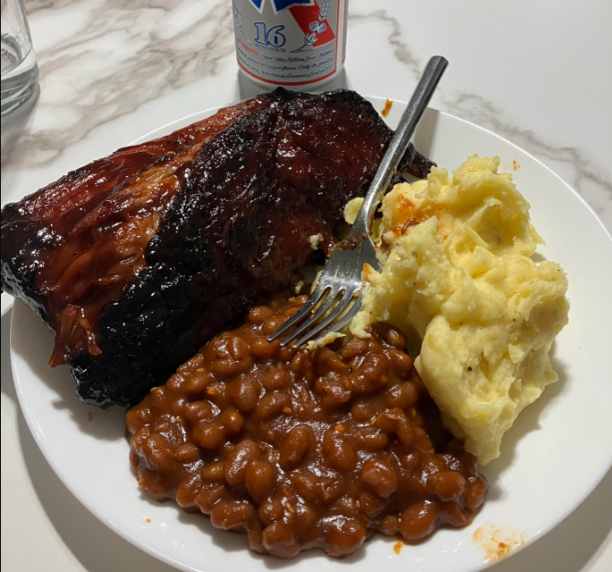 A plate with baked beans, mashed potatoes, and barbecue ribs, with a fork on the plate and a can of beer in the background on a marble surface.