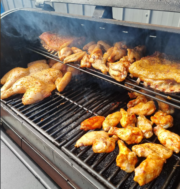 A smoker filled with various pieces of seasoned chicken, including whole chickens, wings, and thighs, cooking on both upper and lower racks with smoke visible.