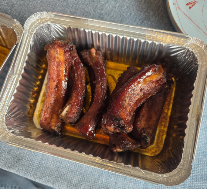 Five cooked pork ribs with a glazed coating sit in a rectangular aluminum takeout container on a gray surface.