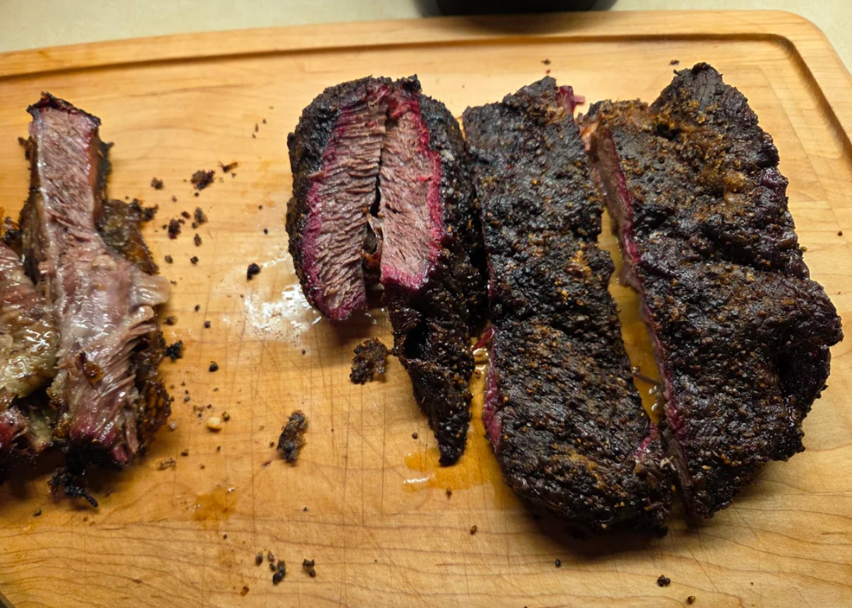 Sliced smoked brisket with a dark, seasoned bark sits on a wooden cutting board. Juices and bits of brisket are visible around the meat.