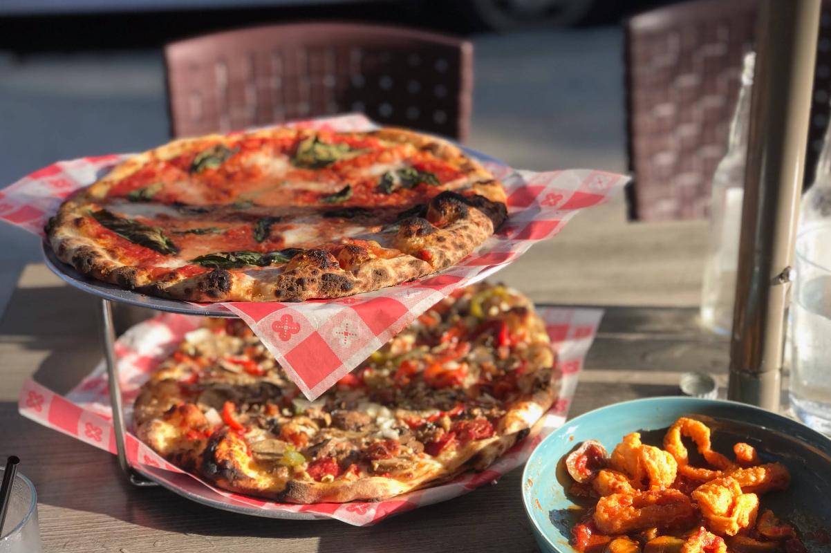 Two pizzas on a tiered stand sit on a table outdoors, one topped with basil and the other with vegetables—perfect for those craving a quality slice of pizza for cheap. Nearby, find a blue bowl of saucy wings or bites, clear glasses, and bottled water.