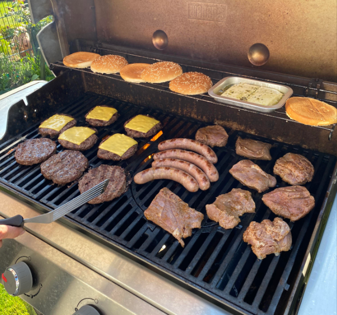 A barbecue grill with burger patties, sausages, meat, cheeseburgers, and buns. A spatula is flipping food, and a tray of garlic butter is heating. The setting appears to be an outdoor cookout.