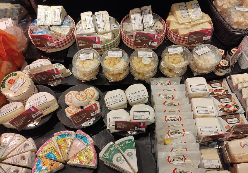 A variety of packaged cheeses from different countries, including France and Denmark, displayed on a store shelf with visible labels and assorted shapes.