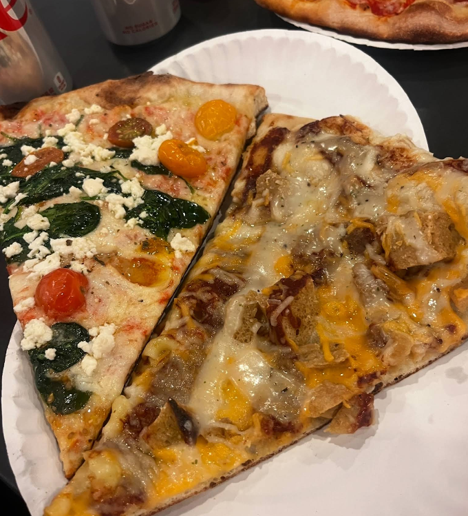 Two large pizza slices on a white paper plate: one with spinach, cherry tomatoes, and feta; the other with melted cheese, barbecue chicken, and onions—a quality slice of pizza for cheap. Part of another pizza and soda cans appear in the background.