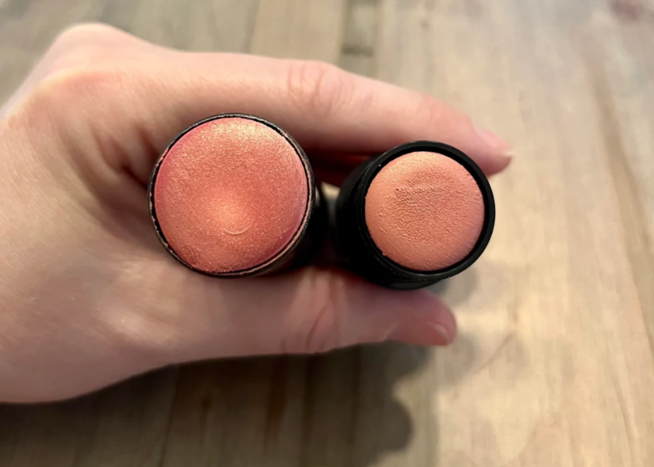 A hand holds two round, peach-colored makeup sticks with slightly different shades, shown from above on a wooden surface—an image reminiscent of photos of shrinking products, highlighting subtle variations in size and hue.