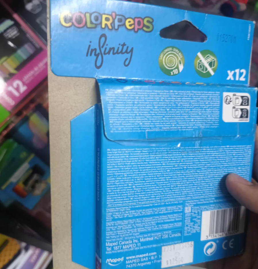 A close-up of a hand holding a blue box of Color'Peps Infinity colored pencils, 12 pack, with shelves displaying similar art supplies and photos of shrinking products visible in the background.