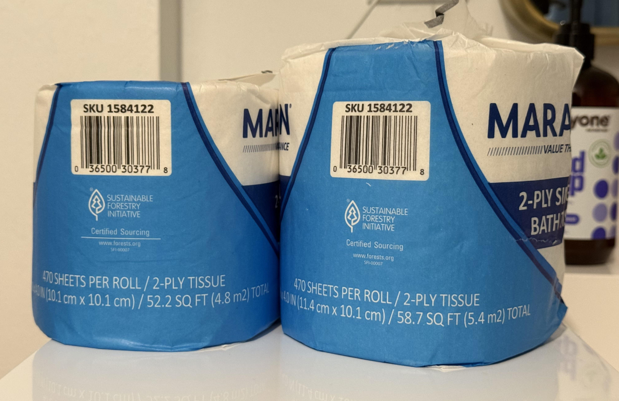 Two rolls of blue and white toilet paper with barcodes on top, showing product details and Sustainable Forestry Initiative certification—capturing the trend seen in many photos of shrinking products. A brown bottle is blurred in the background.