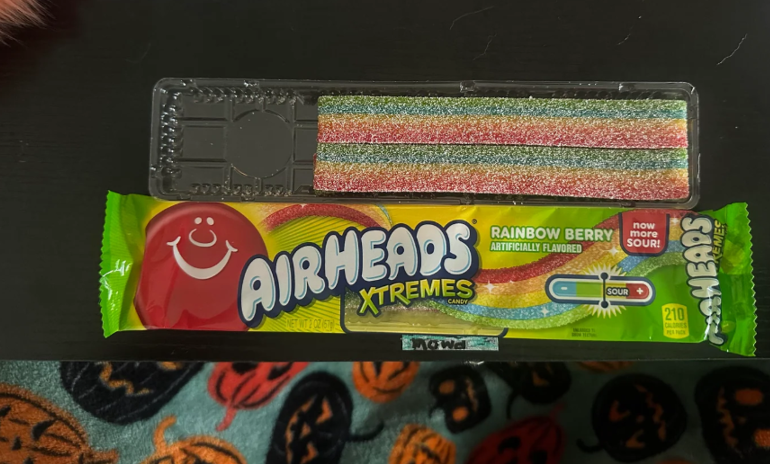 A pack of Airheads Xtremes Rainbow Berry sour candy, with one multicolored candy strip in a clear tray above the opened wrapper. The Halloween-themed blanket is partly visible underneath, evoking those familiar photos of shrinking products that highlight how much smaller favorite treats have become over time.