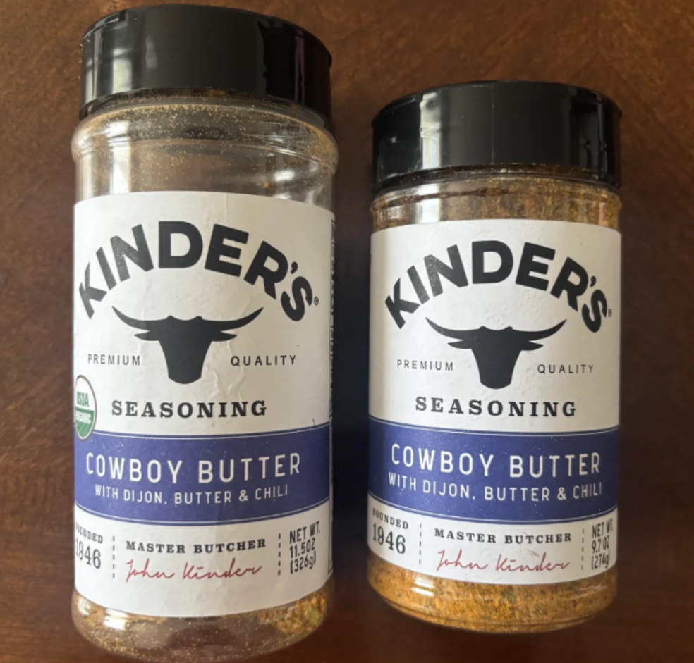 Two jars of Kinder's Cowboy Butter Seasoning with Dijon, butter, and chili stand side by side on a wooden surface. Both jars have black lids and white labels featuring a bull head logo and John Kinder’s signature. Popular in recent photos of shrinking products, these generous jar sizes remind you what real value looks like.