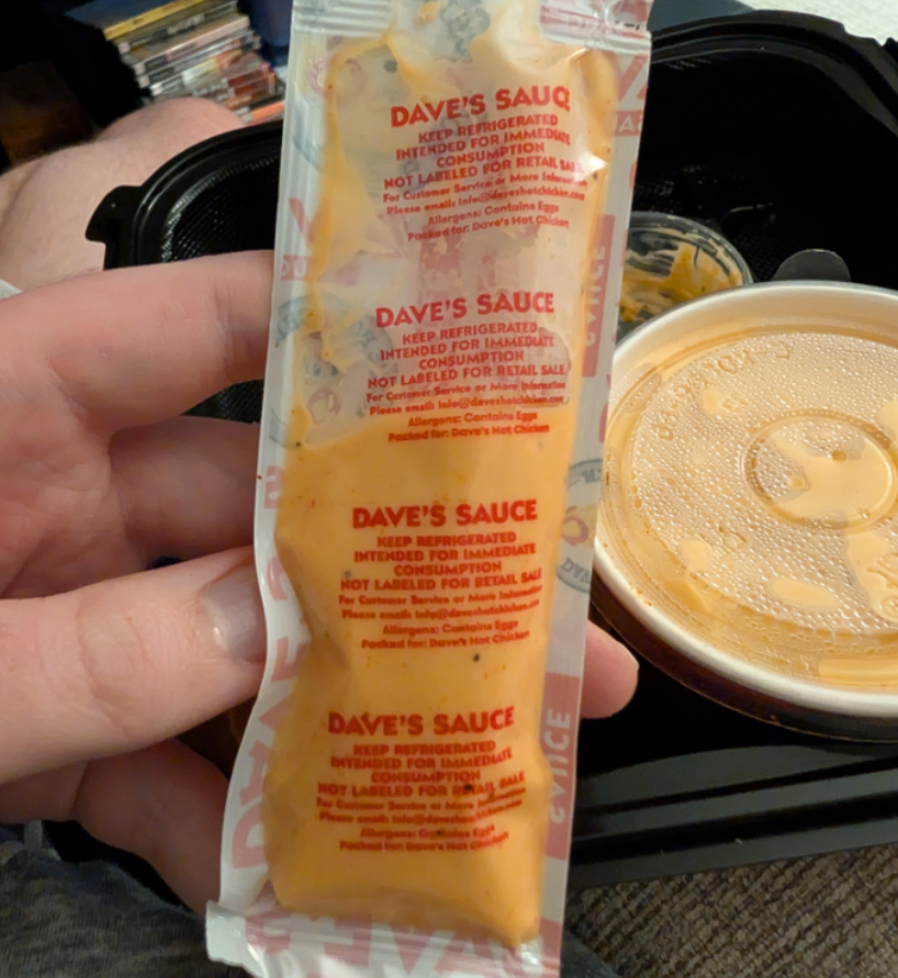 A hand holds a packet labeled “Dave’s Sauce” in red text. In the background are a takeout container with food, a lidded cup with sauce, and part of a person’s leg—capturing one of those moments you notice from photos of shrinking products.