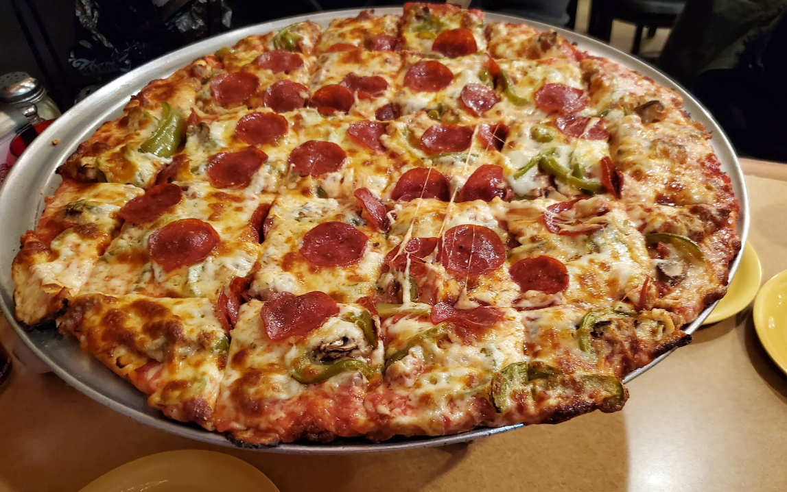 A large rectangular pizza cut into square pieces, topped with melted cheese, pepperoni, green bell peppers, and mushrooms—served on a round metal tray so you can enjoy a quality slice of pizza for cheap at the restaurant table.