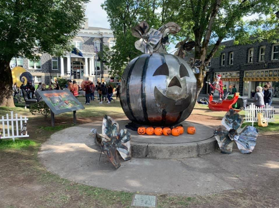 Large metallic jack-o’-lantern sculpture with three metal flowers, surrounded by real pumpkins, in a park with people, a white house, trees, and picket fences. Festive Halloween atmosphere adds to the fall festival charm.