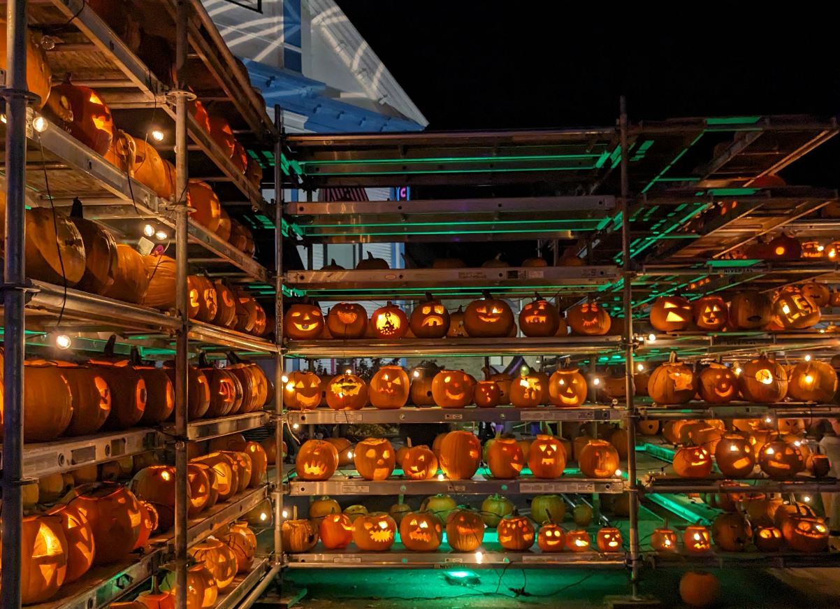 Rows of carved, illuminated jack-o’-lanterns are displayed on metal shelves at night, each with unique faces and designs, creating a festive Halloween atmosphere that captures the spirit of the season.