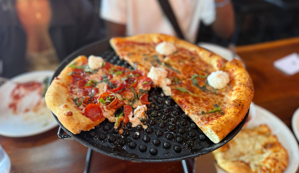A pizza on a black serving tray, partially eaten, with pepperoni and cheese slices offers a quality slice of pizza for cheap. Dollops of ricotta cheese top a few slices. Plates with leftover pizza crusts appear in the background.
