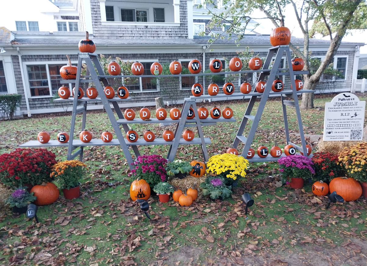 A festive halloween display of pumpkins painted with letters, arranged on ladders and surrounded by colorful flowers and pumpkins. A white house stands in the background, while a sign about a pumpkin jumble is positioned to the right.