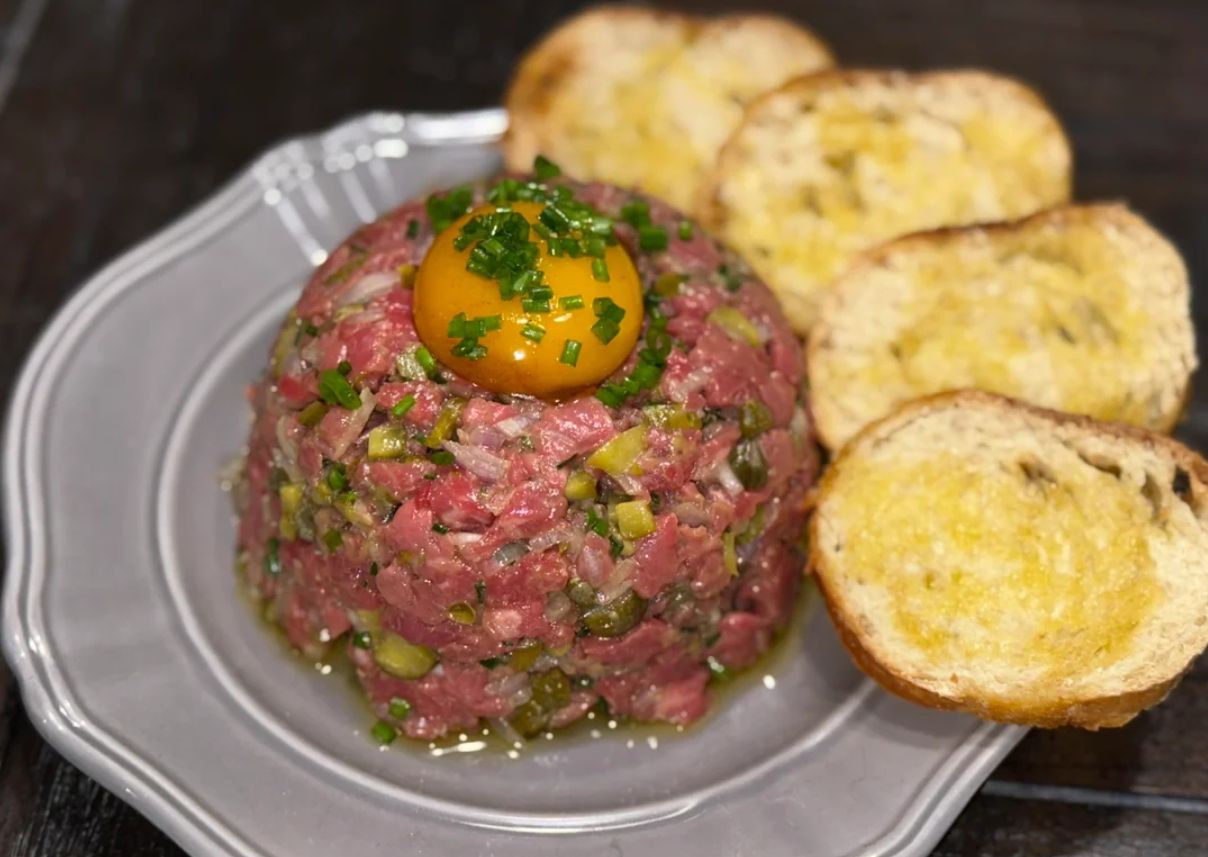 A mound of beef tartare topped with an egg yolk and chopped chives, served with slices of toasted bread on a gray plate.