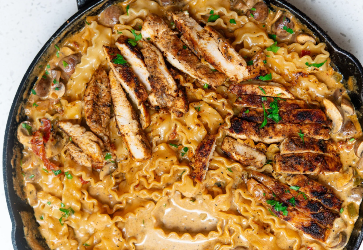 A skillet of creamy pasta with ruffled edges, topped with sliced grilled chicken and garnished with chopped parsley.