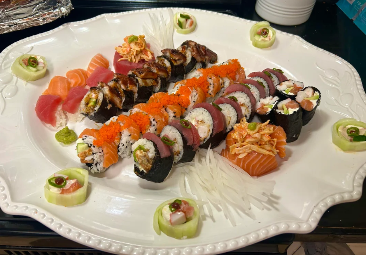 A platter of assorted sushi rolls, sashimi slices, and garnishes, arranged on a white plate with wasabi, pickled ginger, cucumber flowers, and shredded daikon.