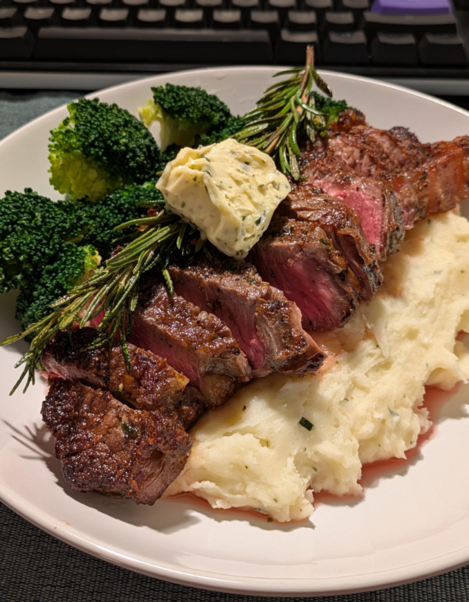 Sliced steak on mashed potatoes, topped with herb butter and a sprig of rosemary, served with steamed broccoli. A keyboard is visible in the background.