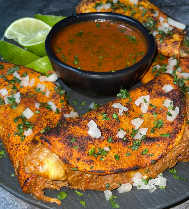 Three crispy birria tacos filled with meat and melted cheese, topped with chopped onions and cilantro, served with lime wedges and a bowl of rich, red consommé for dipping.
