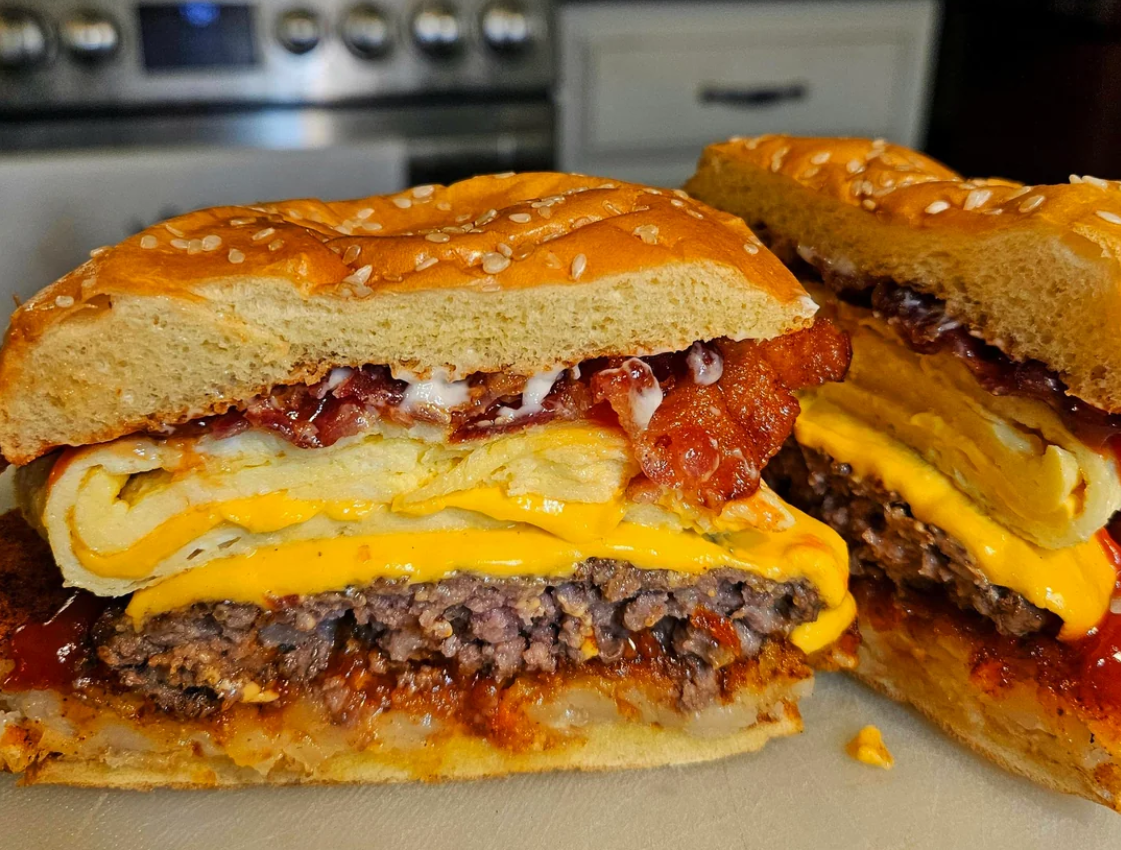 A close-up of a sandwich cut in half, showing layers of ground beef patty, cheddar cheese, folded egg, bacon, and sauce between sesame seed buns, with a kitchen stove in the background.